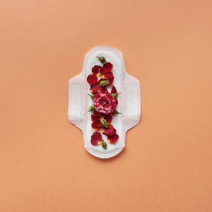 A sanitary pad adorned with red rose petals on a peach background, symbolizing menstruation and femininity.